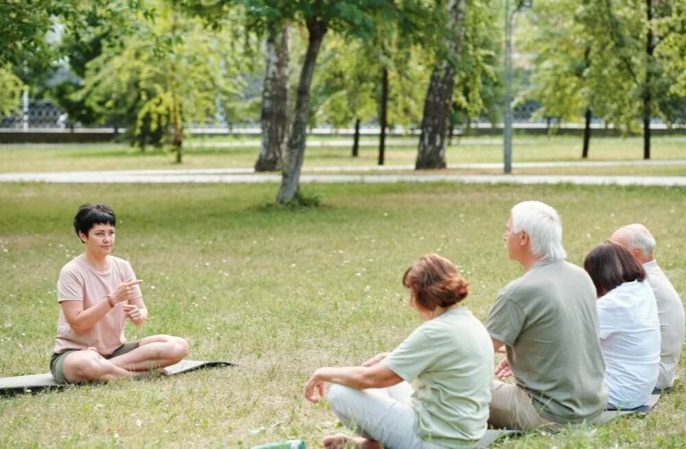 Person leading meditation session with a small group of people seated on the grass in a park. - Home Instead