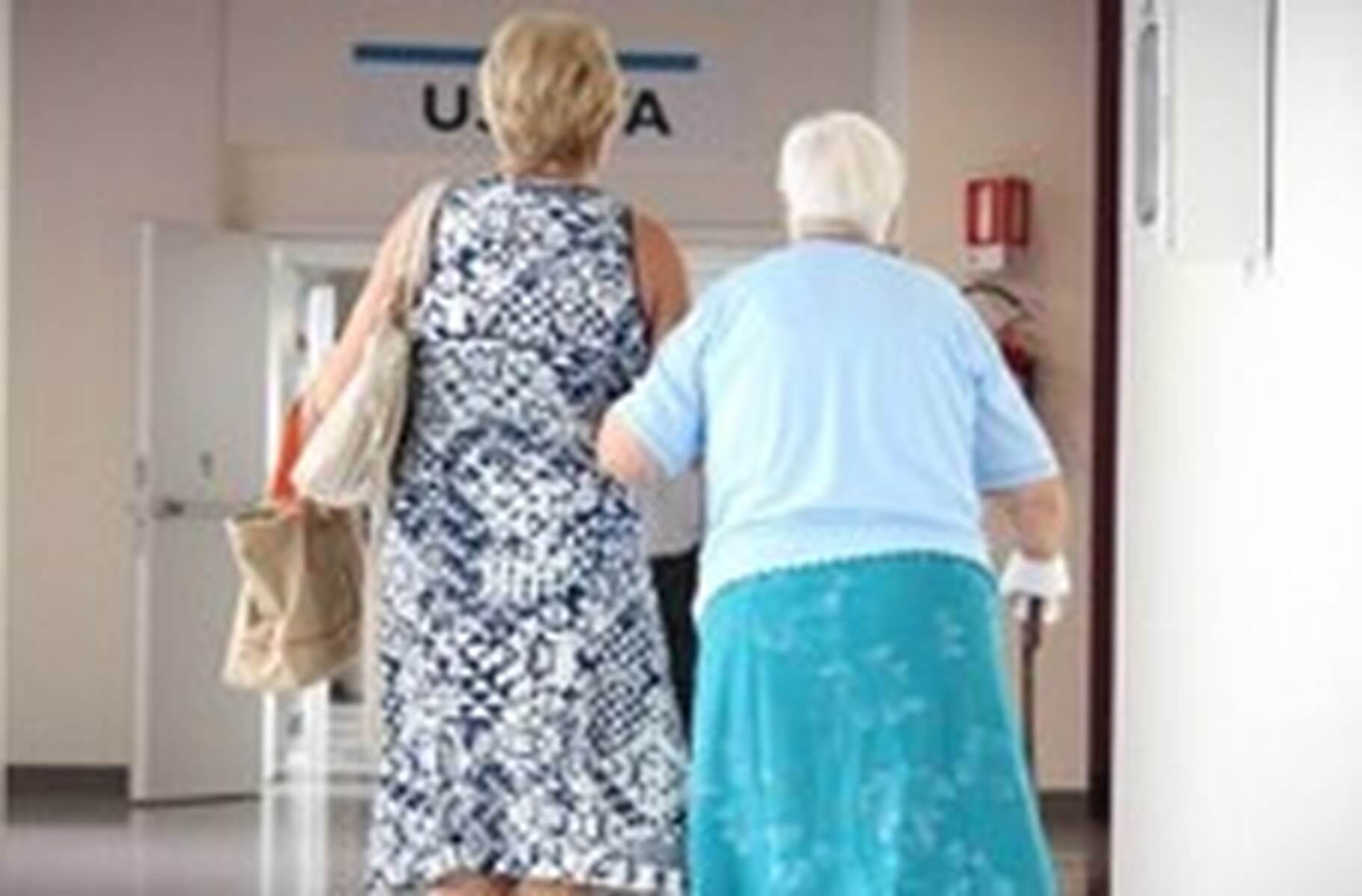 Two elderly people walking down a hallway, one using a cane, with a sign marked "U.S.L.A" above an open door. - Home Instead