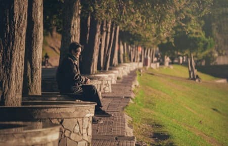 A person sitting alone on a stone bench along a tree-lined pathway in a park during the daytime. - Home Instead