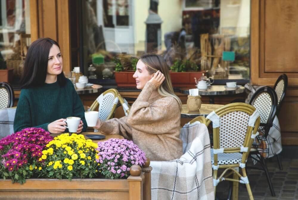 Two women enjoying coffee and chatting at an outdoor café, with colorful flowers in the foreground. - Home Instead