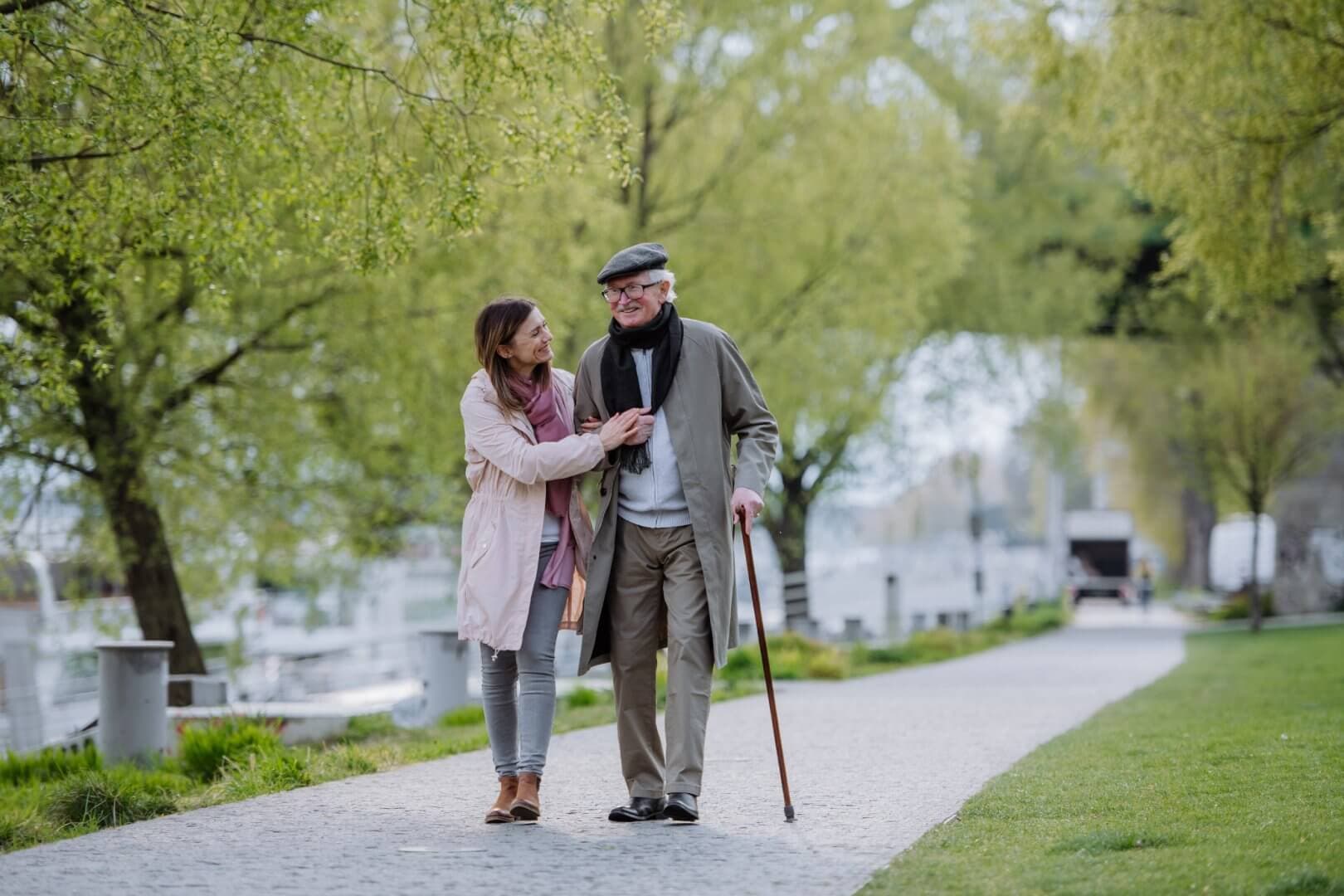 An elderly man with a cane and a younger woman stroll together on a tree-lined path, enjoying a pleasant conversation. - Home Instead