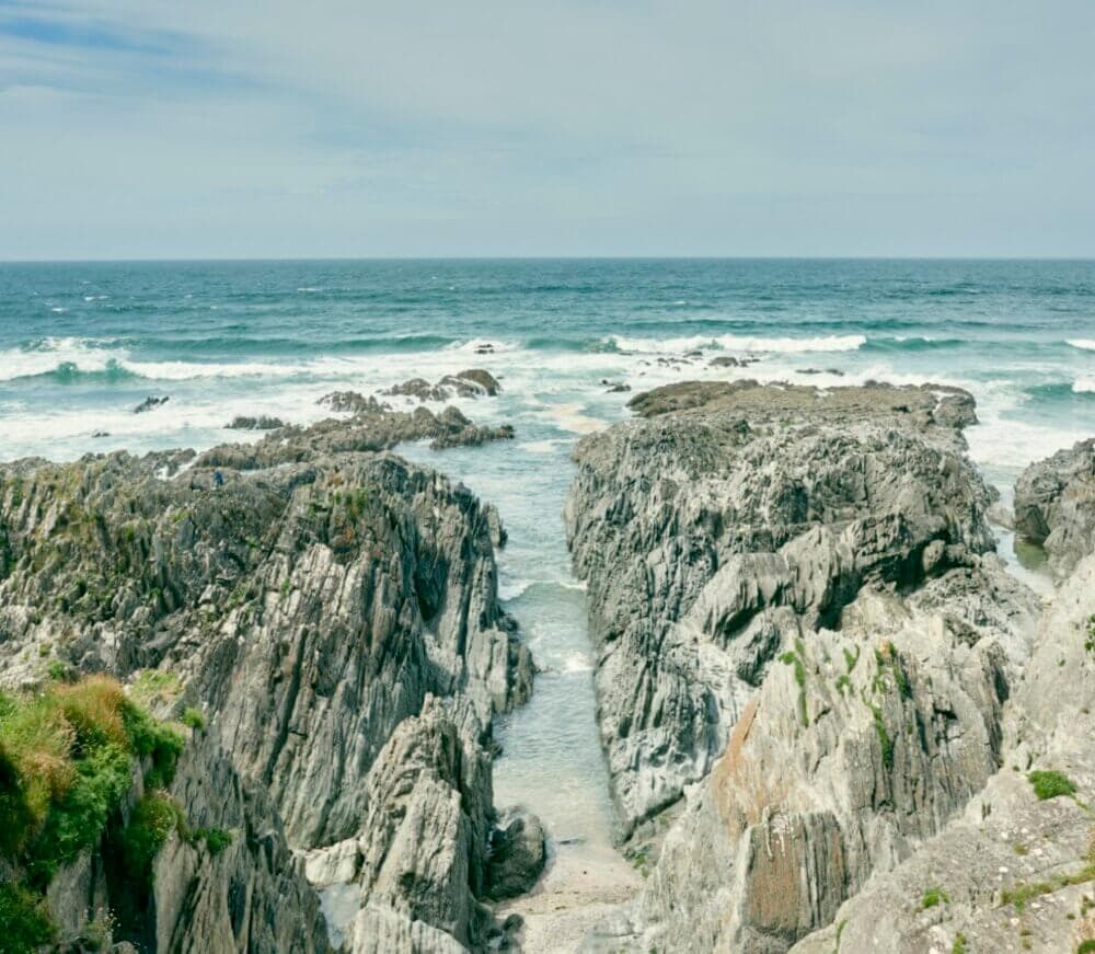 Rocky shoreline with steep cliffs and crashing ocean waves under a partly cloudy sky. - Home Instead