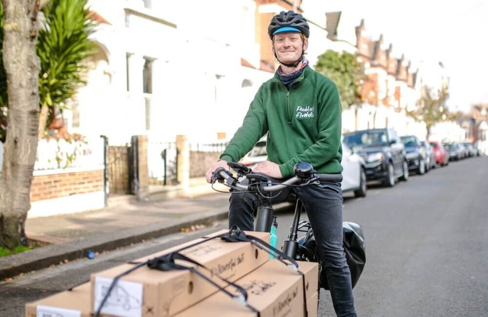 A cyclist wearing a helmet and green jacket is transporting boxes on a cargo bike along a residential street. - Home Instead