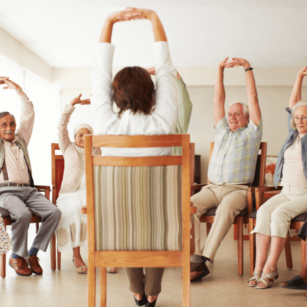 A group of elderly people sitting in chairs, stretching their arms up during a group exercise class. - Home Instead