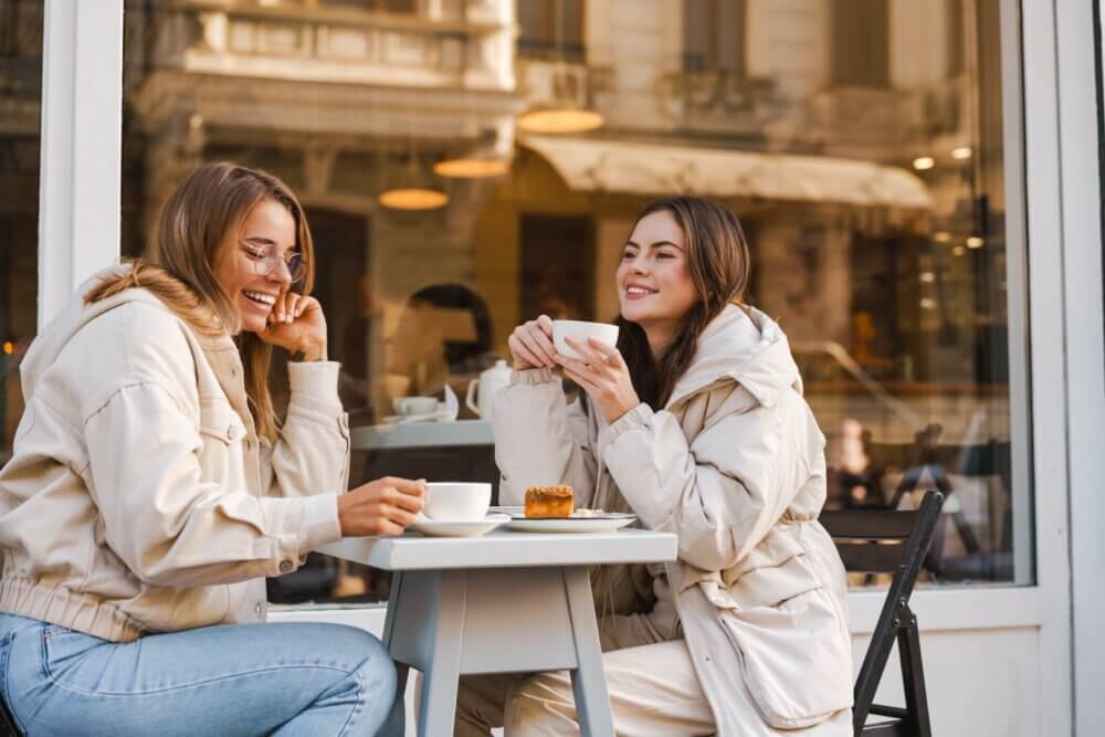 Two women in winter jackets enjoy coffee and pastries at an outdoor cafe, smiling and chatting. - Home Instead