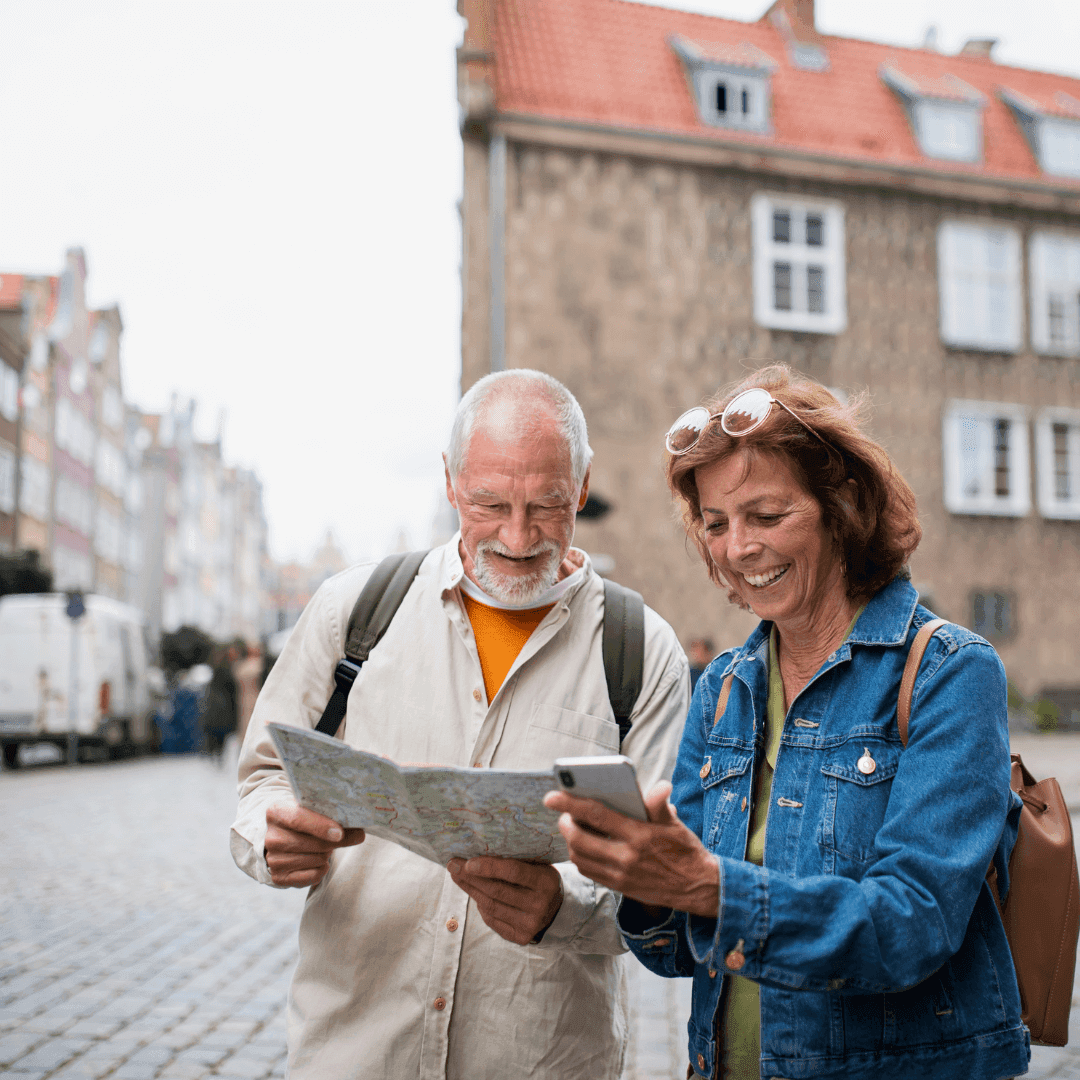 An older couple, one holding a map and the other a phone, smile while sightseeing on a cobblestone street in a European city. - Home Instead
