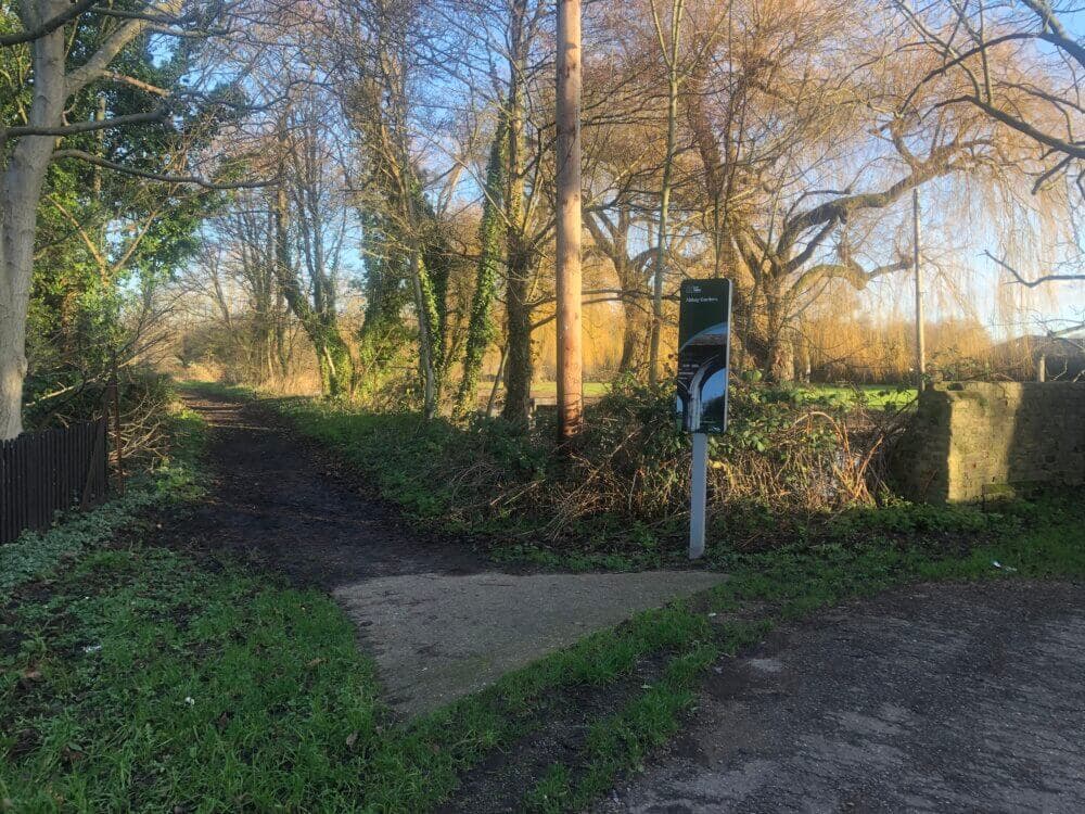 A pathway surrounded by leafless trees and greenery on a sunny day, with a signpost on the right of the path. - Home Instead