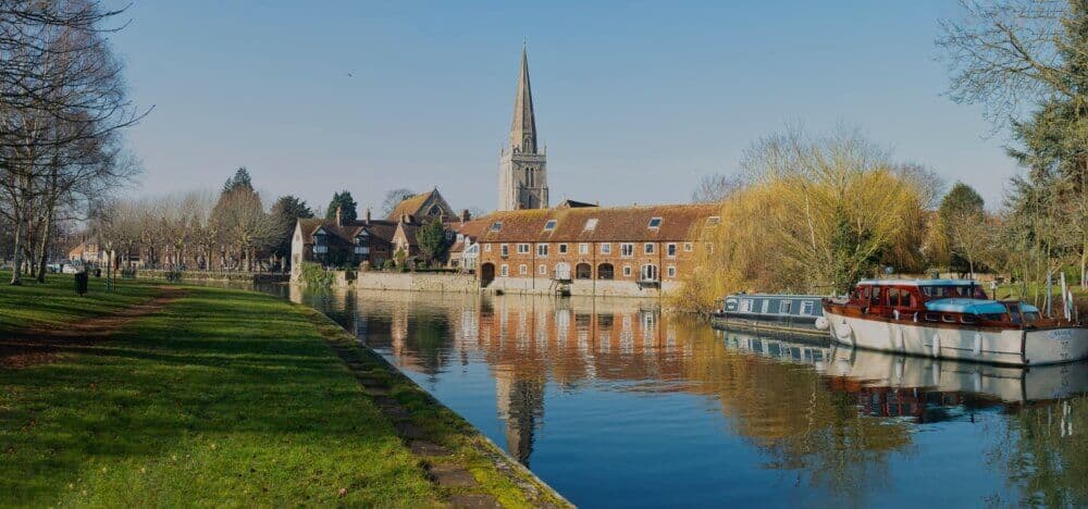 A peaceful riverside scene with boats, historic buildings, and a church spire under a clear blue sky. - Home Instead