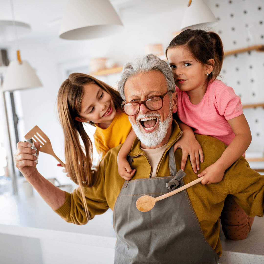 An elderly man with gray hair and glasses cheerfully cooking with two young girls in a bright kitchen. - Home Instead