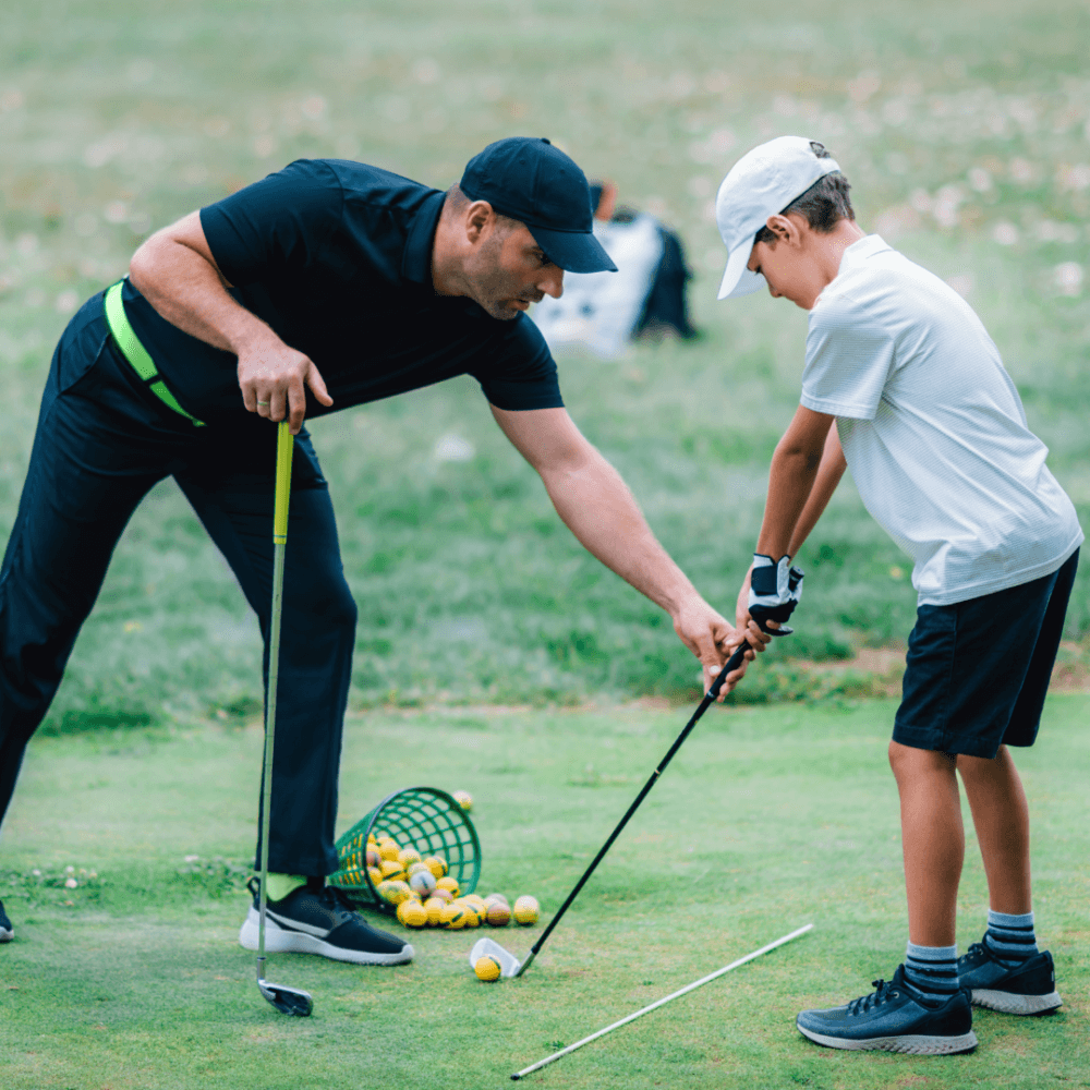 An adult helps a child with his golf swing, guiding his grip. They are on a grassy area with scattered yellow golf balls. - Home Instead