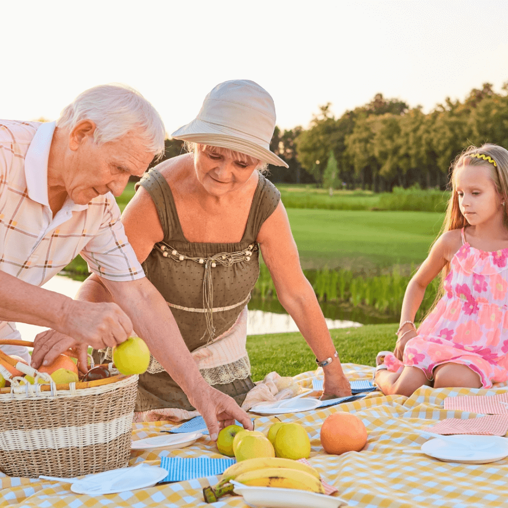 A man and woman unpack a picnic basket on a blanket with fruit; a young girl in a pink dress is sitting nearby. - Home Instead