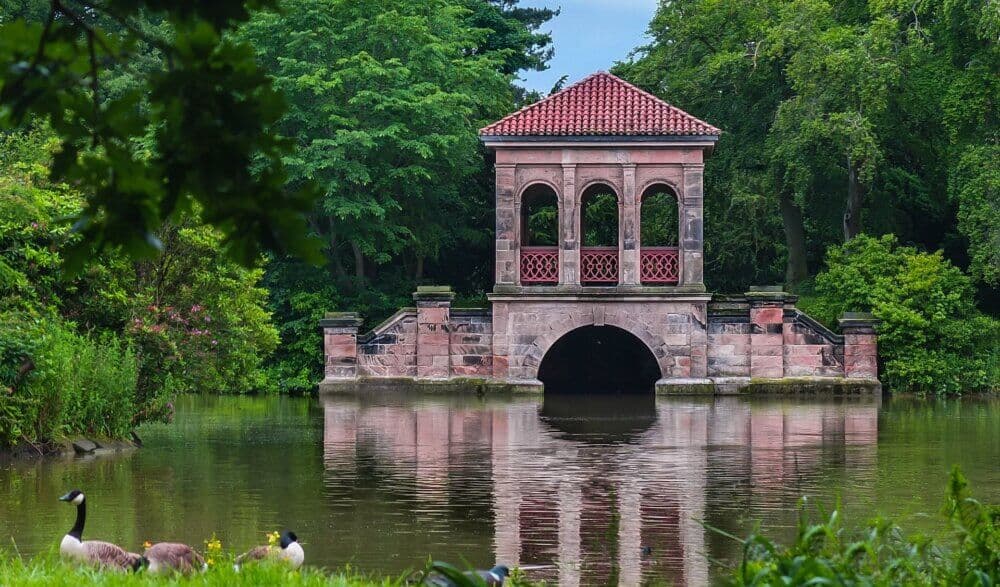 Stone pavilion with arched openings, red-tiled roof, surrounded by lush greenery, reflecting in a calm pond. - Home Instead