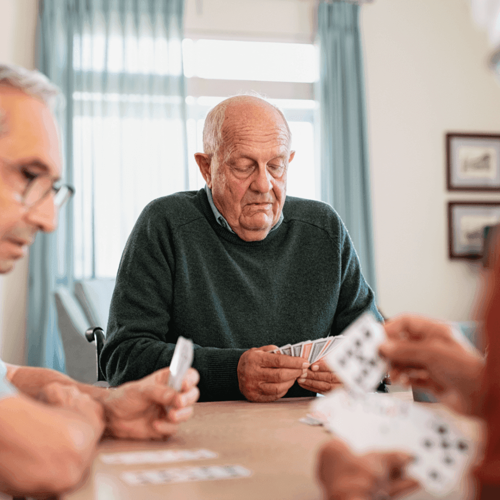 Three elderly individuals play cards together at a table in a bright room with curtains and framed pictures in the background. - Home Instead