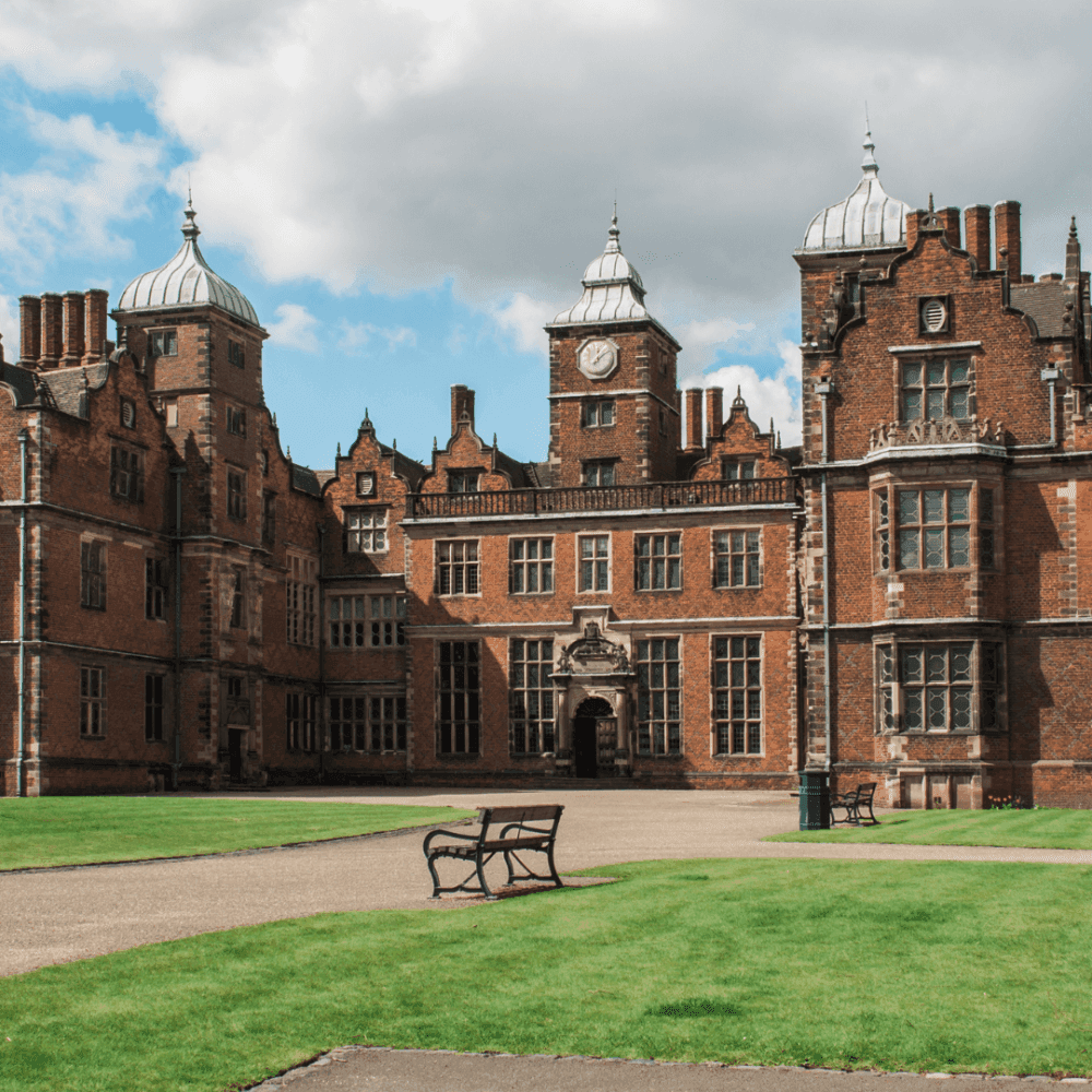 Historical brick building with towers, a clock, and a paved courtyard with benches and grass, under a partly cloudy sky. - Home Instead