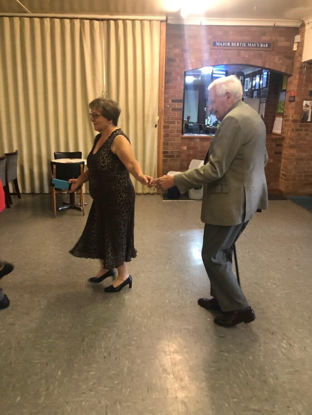 Two elderly individuals are dancing together in a room with a brick wall and a small bar in the background. - Home Instead