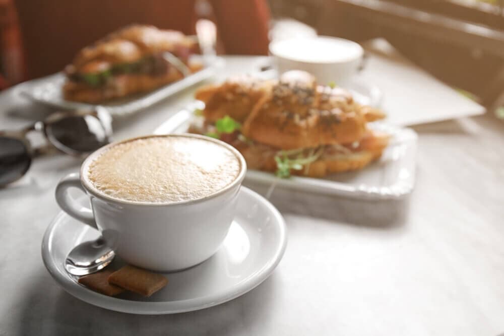 A frothy cappuccino in a white cup with croissant sandwiches on plates in the background. - Home Instead