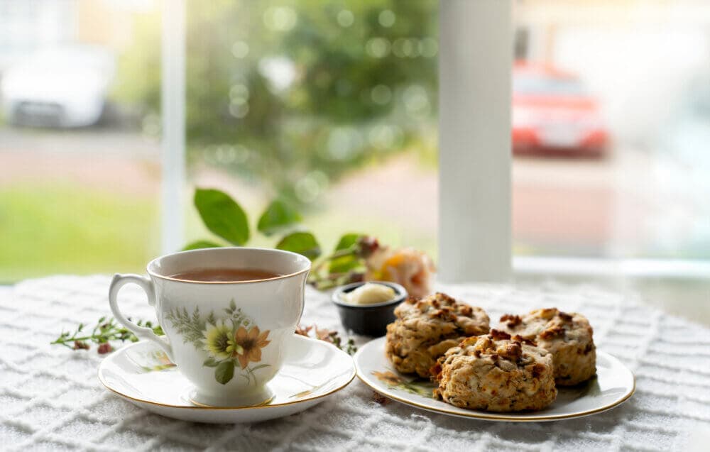 A cup of tea with floral design sits next to a plate of scones with butter on a table near a window. - Home Instead