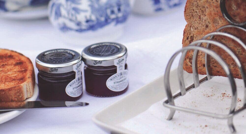 Two jars of strawberry jam next to a knife, a toasted bread slice, and a bread holder on a white tablecloth. - Home Instead
