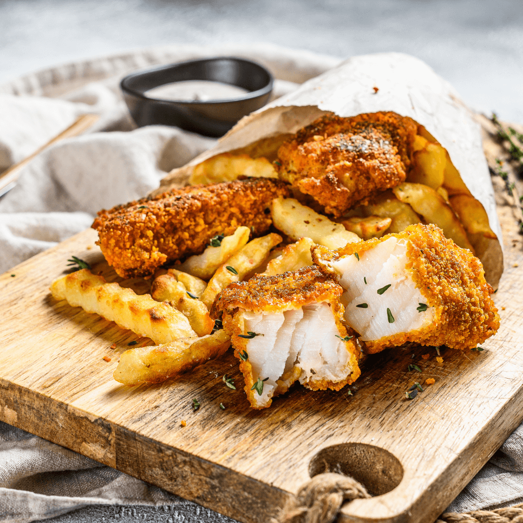Crispy fried fish and golden fries spilling out of a paper bag onto a wooden board, with a small dipping sauce in the background. - Home Instead