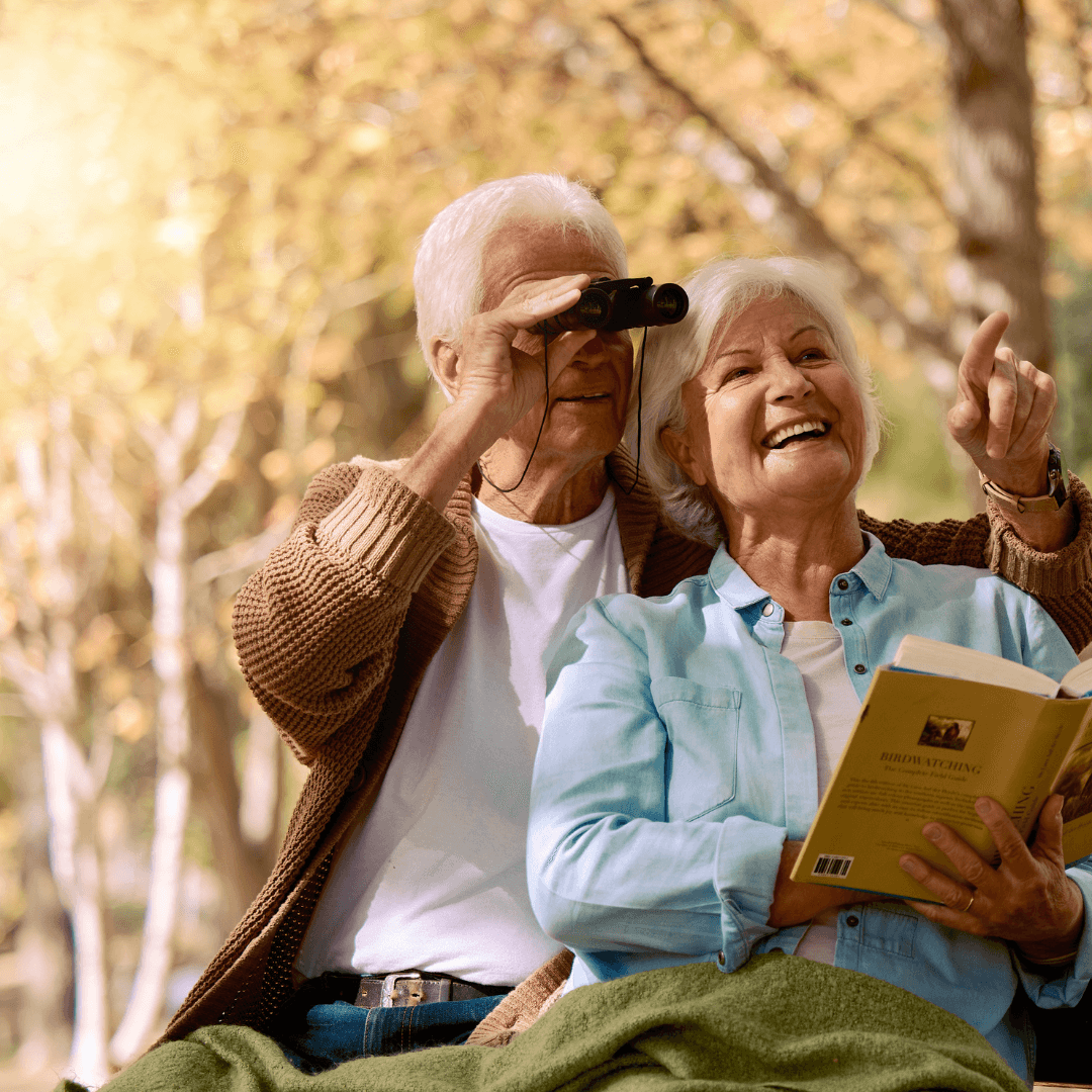 Elderly couple enjoying birdwatching; the man uses binoculars while the woman smiles and points, holding a book. - Home Instead