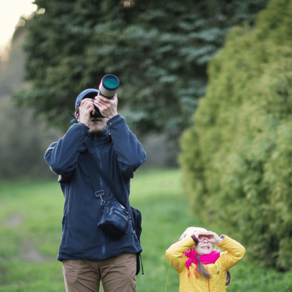 An adult and child are birdwatching outdoors, the adult uses binoculars while the child shields eyes with hands. - Home Instead
