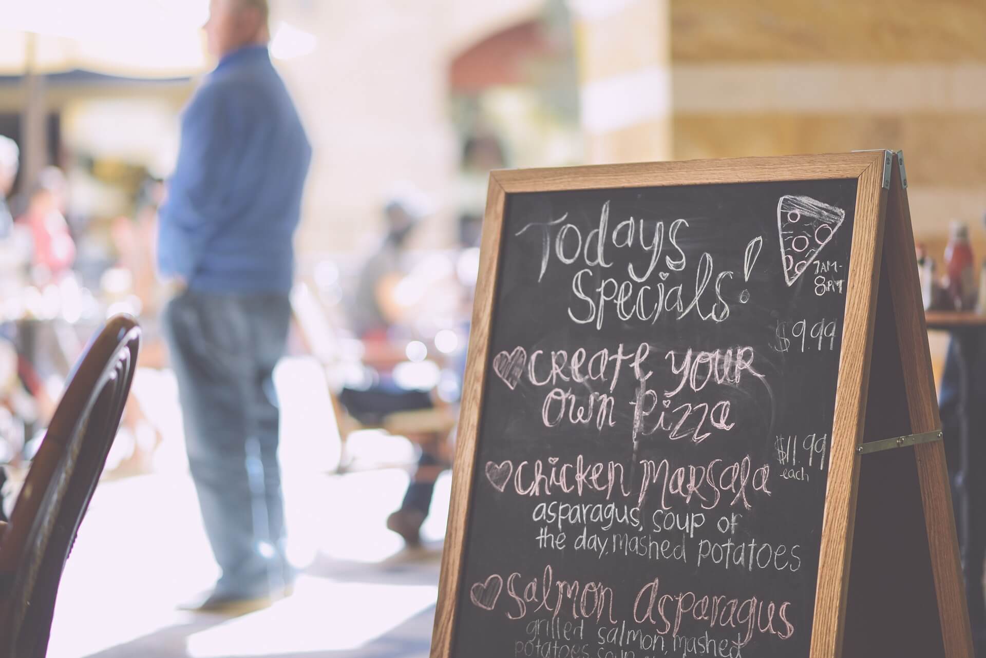 Chalkboard menu at a cafe offering daily specials with a blurred background of customers and outdoor seating. - Home Instead