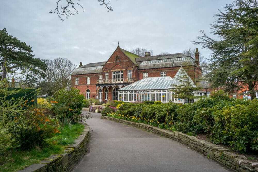 A path leads to a historic brick building with a glass conservatory, surrounded by gardens and greenery on a cloudy day. - Home Instead