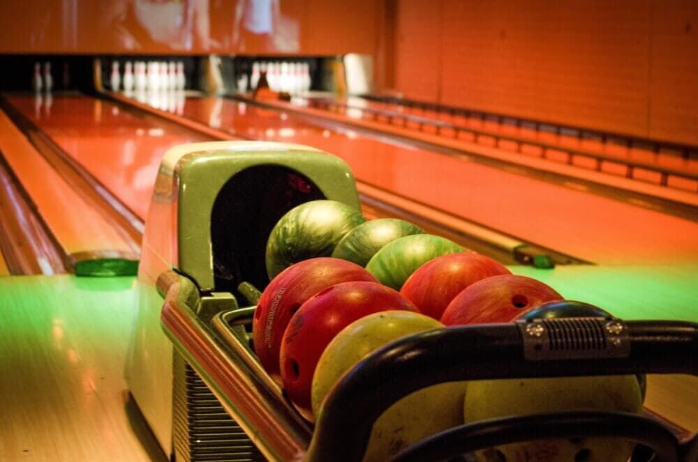 Close-up of a bowling ball rack with green and red balls on it, next to a bowling lane. Pins are visible in the distance. - Home Instead