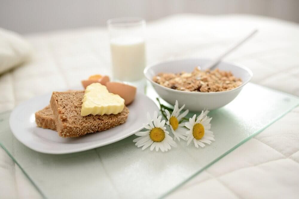 Breakfast with bread, butter, eggs, a bowl of cereal, and a glass of milk on a tray with daisies. - Home Instead
