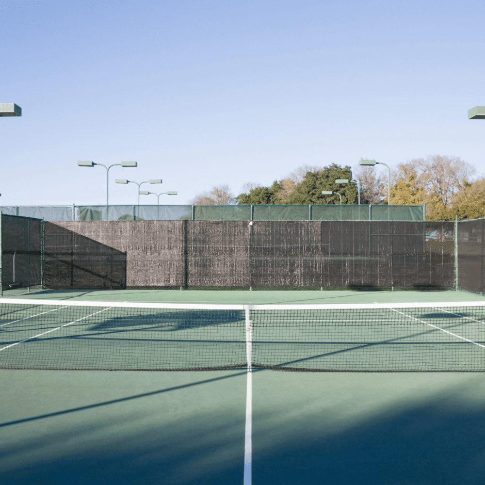 Empty outdoor tennis court with a green surface and surrounding high fences on a clear, sunny day. - Home Instead