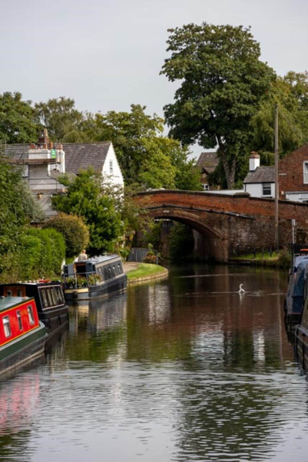 A narrow canal with houseboats, tree-lined banks, and a red brick bridge in the background on a cloudy day. - Home Instead