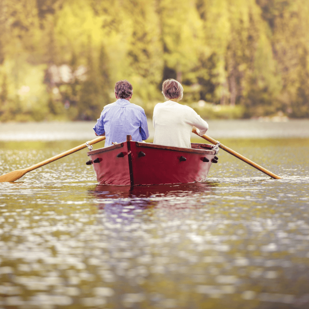 Two people rowing a red boat on a calm lake surrounded by green trees on a bright day. - Home Instead