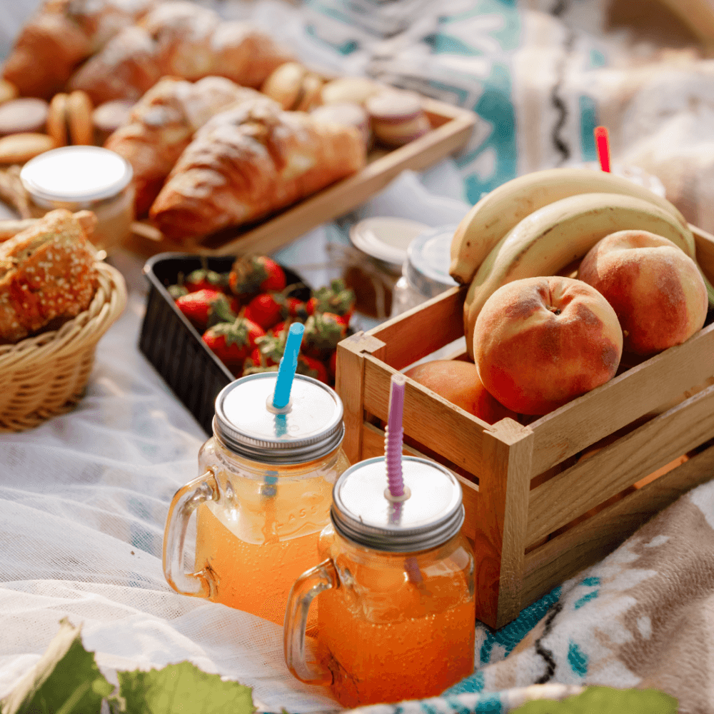 Picnic setup with pastries, strawberries, peaches, bananas, and two mason jar drinks with straws on a blanket. - Home Instead