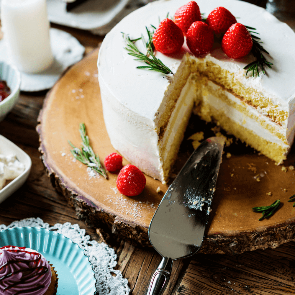 A sliced white cake with strawberries on top, displayed on a wooden board, surrounded by cupcakes and a cake server. - Home Instead