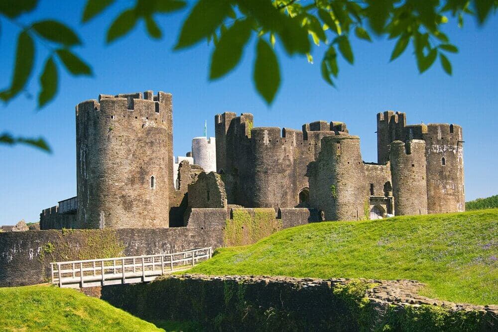 Medieval stone castle with a wooden bridge entrance, surrounded by greenery, under a bright blue sky with leafy branches. - Home Instead