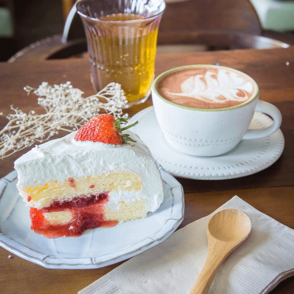 A slice of strawberry shortcake with whipped cream, a cup of latte with art, and a glass of iced tea on a wooden table. - Home Instead