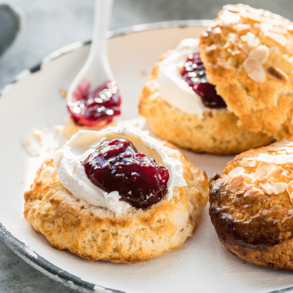 Two scones with cream and red jam, one partially covered and a spoon with jam on a white plate. - Home Instead