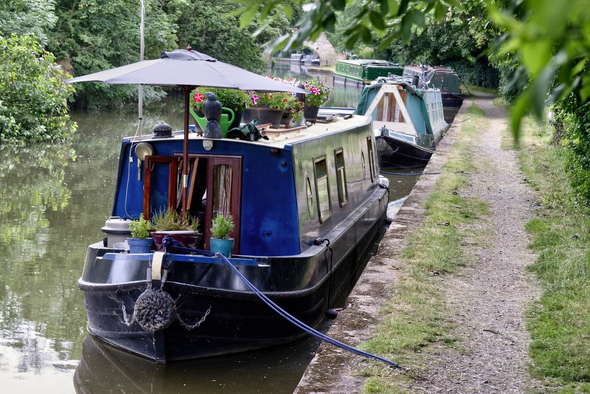 A narrowboat docked along a lush, green canal path with plants and an umbrella adorning the deck. - Home Instead