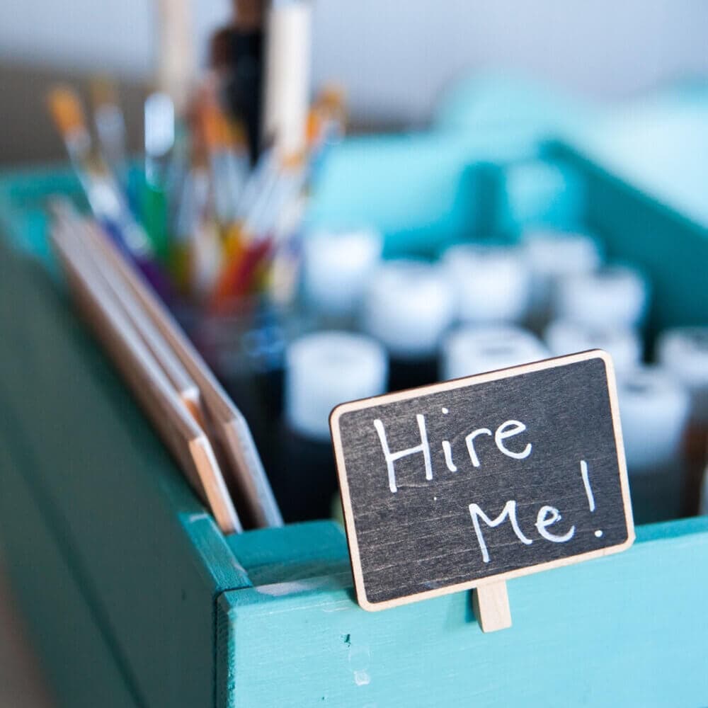 A small chalkboard sign reading "Hire Me!" is placed on a teal wooden box filled with paint bottles and paintbrushes. - Home Instead