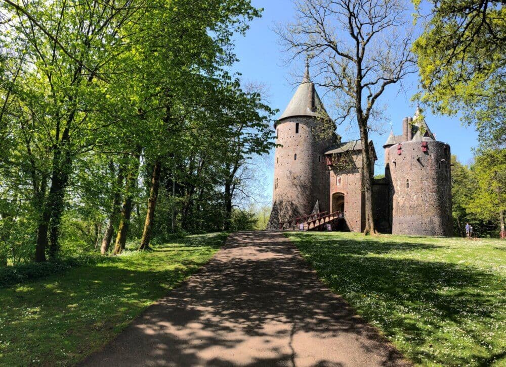 A path leading to an old stone castle with turrets, surrounded by lush green trees and blue sky. - Home Instead