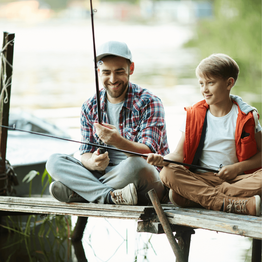 Two people sitting on a dock, fishing and smiling, with water and greenery in the background. - Home Instead