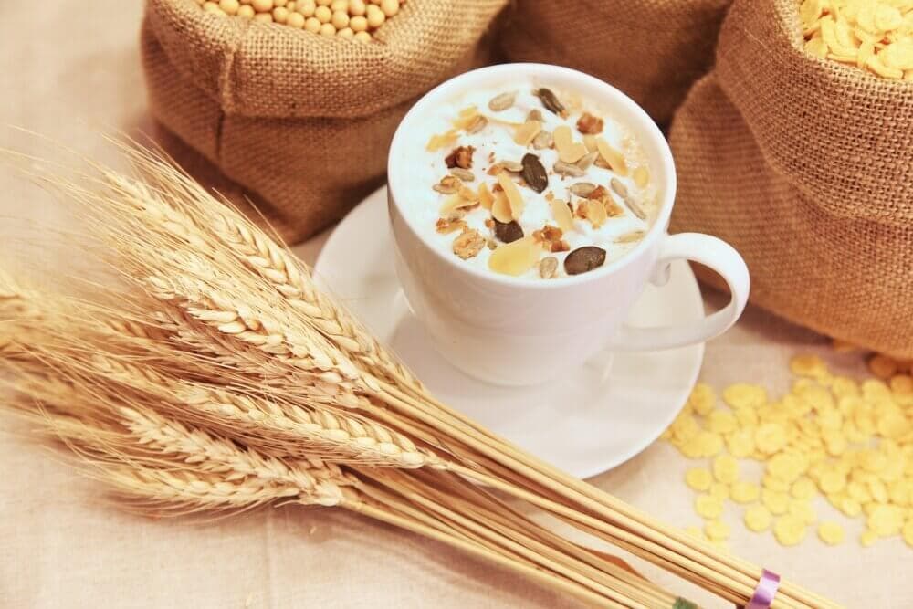 A white cup filled with milk and cereal, surrounded by wheat stalks and burlap sacks of grains. - Home Instead