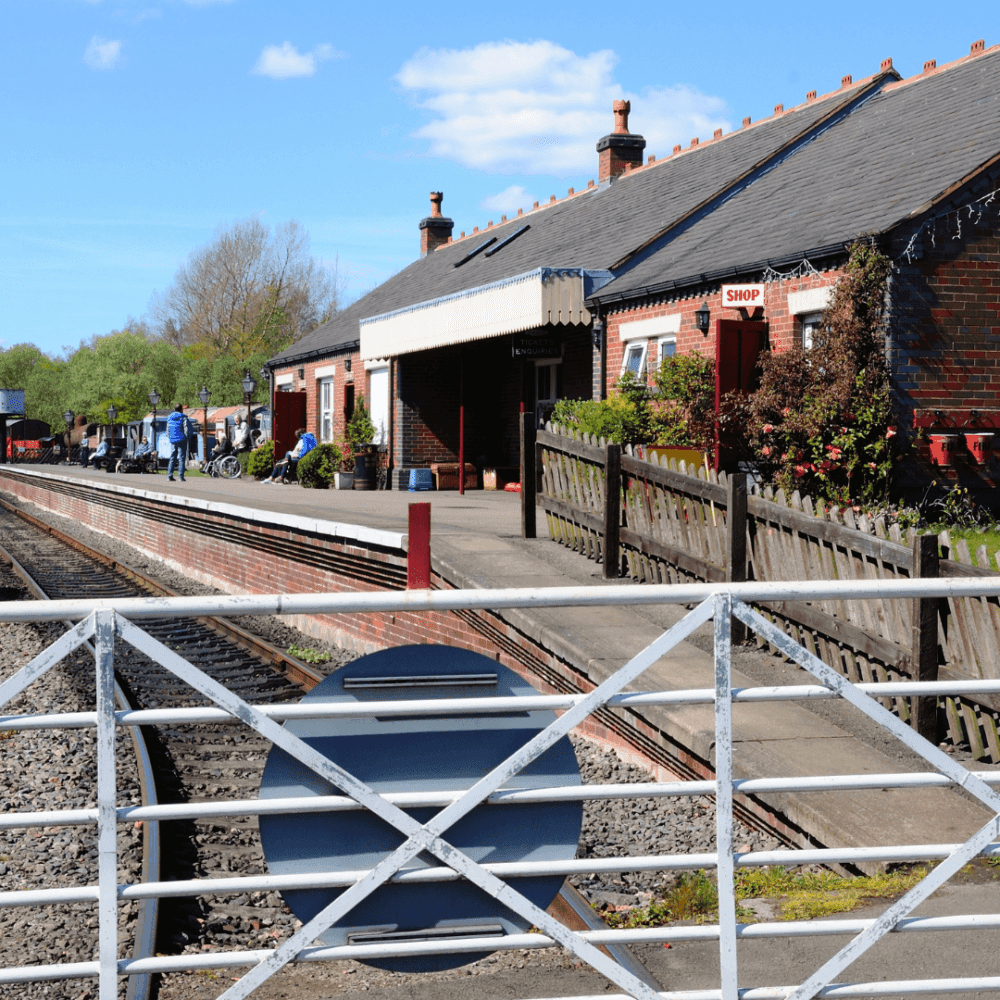 A vintage railway station with brick buildings, platform, shop, and tracks, viewed through a white metal gate. - Home Instead