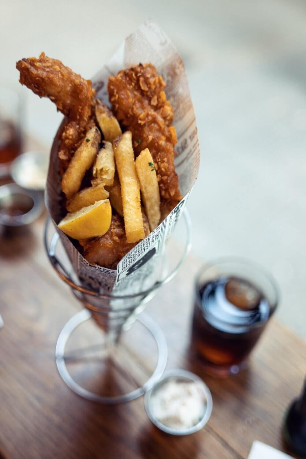 A cone of crispy fish and chips with a lemon wedge on a table next to drinks and dipping sauce. - Home Instead