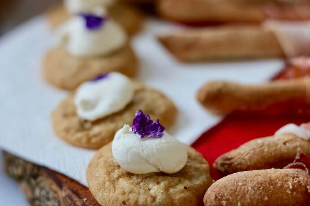 Close-up of cookies topped with dollops of cream and purple flower petals on a wooden surface with other pastries blurred in the background. - Home Instead