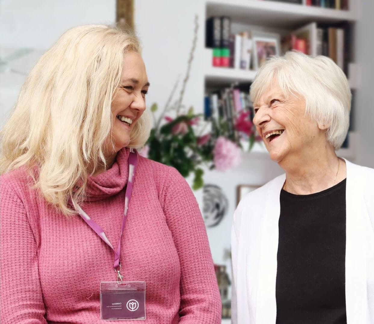 Two women smiling at each other, one wearing a pink sweater with a lanyard, the other in a black top and white cardigan. - Home Instead