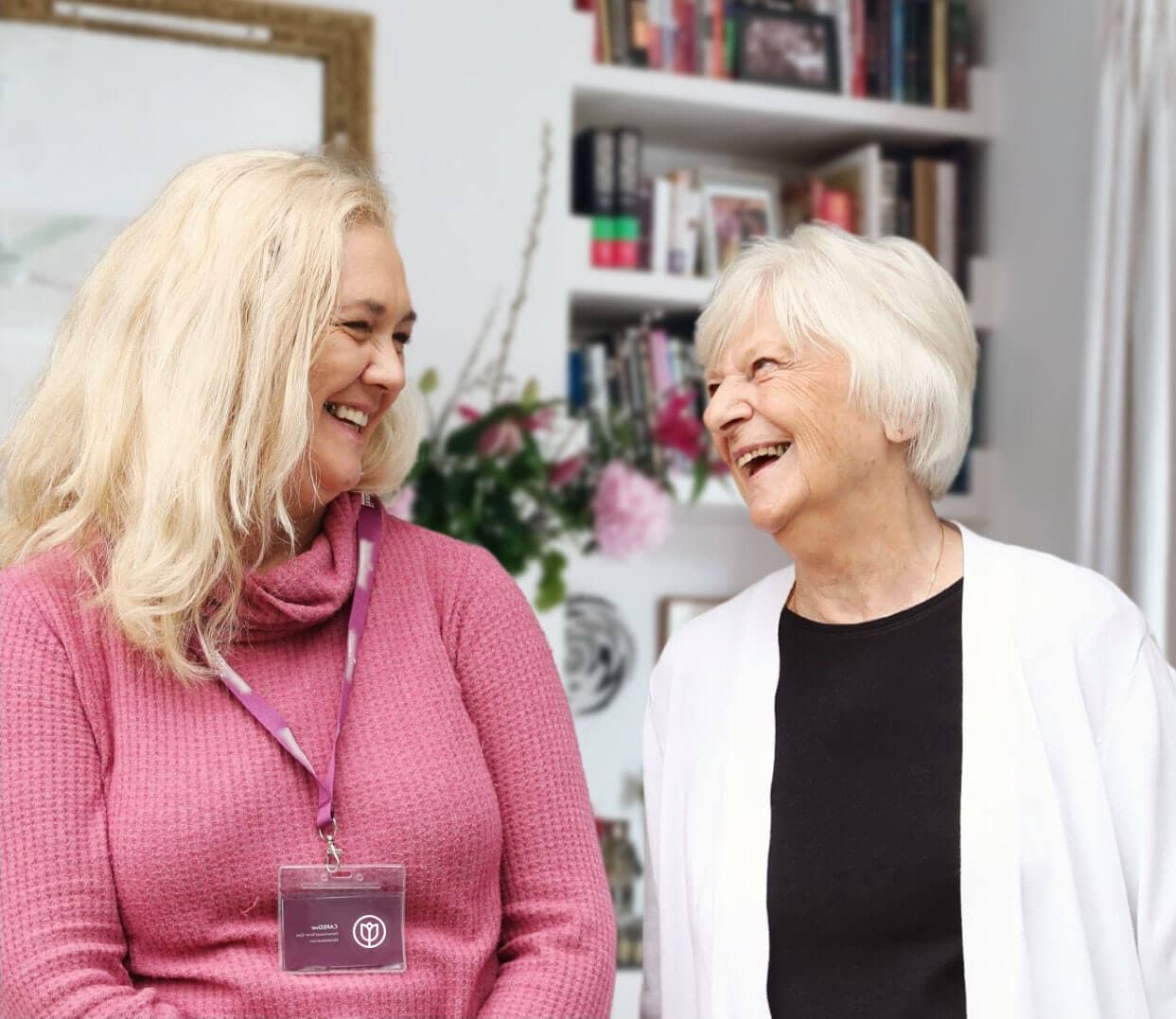 Two women, one younger and one older, smiling and talking in a cozy room with bookshelves and flowers in the background. - Home Instead