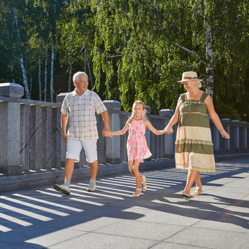 An elderly couple and a young girl walk hand-in-hand on a sunny day across a stone bridge with greenery in the background. - Home Instead
