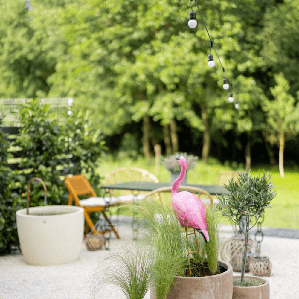 A cozy outdoor garden with a pink flamingo decor, string lights, potted plants, and chairs in a lush green setting. - Home Instead