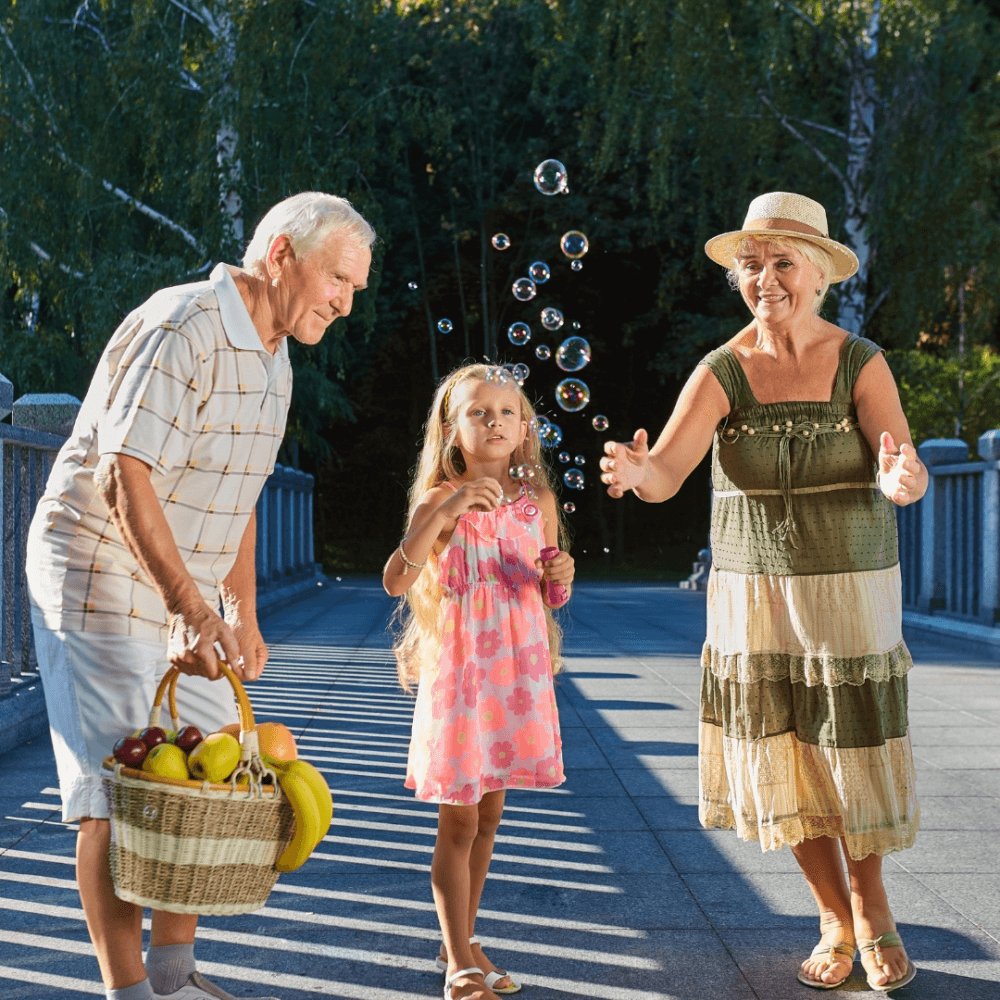 A girl blows bubbles while standing between an elderly man holding a fruit basket and an elderly woman in a hat. - Home Instead